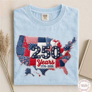 Pode incluir: Camiseta azul clara com um design de patchwork do mapa dos Estados Unidos. O design incorpora tecidos vermelhos, brancos e azuis, com o número "250" e o texto "Years 1776-2026" no centro.