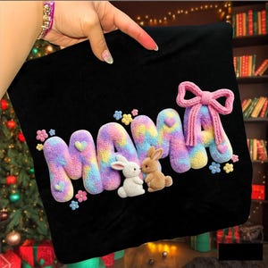 Könnte beinhalten: Schwarzer Stoff mit dem Wort "MAMA" in bunten, aufgeplusterten Buchstaben. Die Buchstaben sind pastellfarben mit Herzen und Blumen. Zwei kleine Hasenfiguren befinden sich in der Mitte. Eine rosa Schleife ziert den letzten Buchstaben.