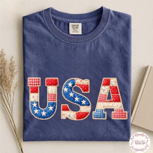 May include: A navy blue t-shirt with the word "USA" appliqu&eacute;d across the front. The letters are made of red, white, and blue fabric in a patchwork design, with the letter "S" featuring white stars on a blue background. The shirt has a crew neck.