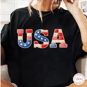May include: Black t-shirt featuring the word "USA" in a patchwork design. The letters are decorated with red, white, and blue patterns, including stars and stripes, reminiscent of the American flag. The shirt is a casual style.