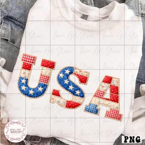 May include: White sweatshirt with the word "USA" in a patchwork design. The letters feature red, white, and blue patterns, including stars and stripes, evoking an American flag theme. The sweatshirt is folded on a denim surface.