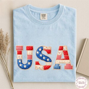 May include: Pale blue Comfort Colors t-shirt featuring the word "USA" in patchwork fabric. The letters are crafted from red, white, and blue patterned fabrics, including stars and stripes. A tag is visible at the neckline.