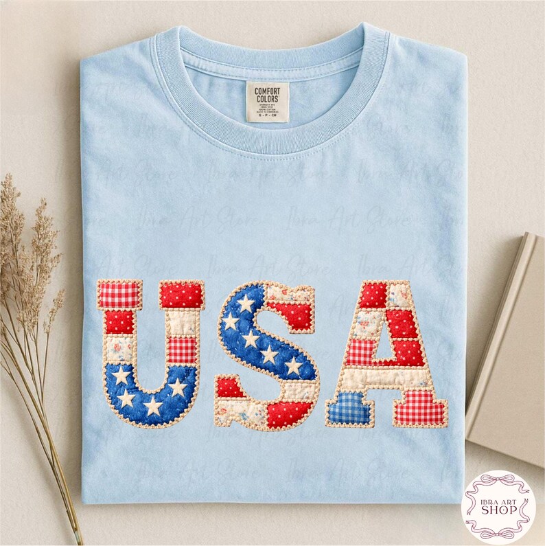 May include: Pale blue Comfort Colors t-shirt featuring the word "USA" in patchwork fabric. The letters are crafted from red, white, and blue patterned fabrics, including stars and stripes. A tag is visible at the neckline.