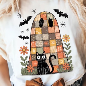 May include: White t-shirt featuring a Halloween-themed graphic. The design includes a patchwork ghost, a black cat, bats, stars, and flowers. The ghost is made of various colorful squares. The cat has large white eyes.
