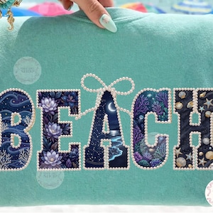 May include: A turquoise sweatshirt with the word "BEACH" in large, pearl-outlined letters. The letters are filled with ocean-themed illustrations, including coral, seashells, and a night sky scene with a moon.