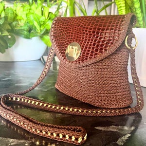 May include: A brown crossbody bag with a textured flap and a gold-colored clasp. The bag features a woven body and a long strap with gold accents. The bag is approximately 8 inches tall and 7 inches wide.
