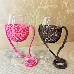 May include: Two wine glasses, each with a crocheted holder and strap. One holder is bright pink, the other is dark brown. The holders are designed to cradle the glass and hang around the neck.