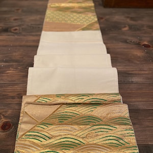 Può includere: Un runner da tavolo decorativo piegato con un centro color crema e estremità a fantasia oro e verde. Le estremità presentano un design a onde con accenti dorati e verdi. Il runner è esposto su una superficie di legno scuro.