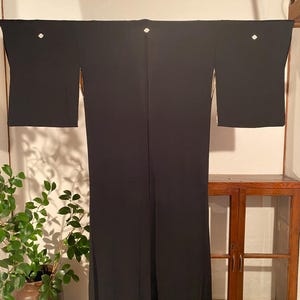 Pode incluir: Um quimono preto com detalhes brancos e um delicado padrão floral. A peça de roupa é exibida pendurada, mostrando todo o seu comprimento e design. As mangas são visíveis, e o tecido parece suave e fluido.