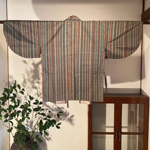 May include: A striped kimono with vertical stripes in shades of blue, red, and beige hangs on a wooden rod. The traditional Japanese garment is displayed against a white wall, showcasing its design and craftsmanship. The sleeves are wide and the fabric appears to be lightweight.