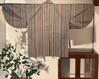 May include: A striped kimono with vertical stripes in shades of blue, red, and beige hangs on a wooden rod. The traditional Japanese garment is displayed against a white wall, showcasing its design and craftsmanship. The sleeves are wide and the fabric appears to be lightweight.