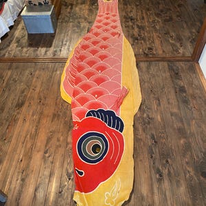 May include: A large, colourful carp streamer, or koinobori, lies on a wooden floor. The fish is primarily red with pink and yellow accents, featuring a detailed eye and scales. The traditional Japanese decoration is used for Children's Day.