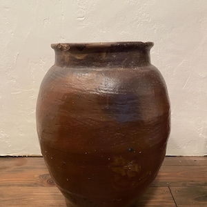 Puede incluir: Una jarra de cerámica marrón antigua y grande con un cuerpo ancho y un borde ligeramente ensanchado. La jarra tiene un aspecto rústico y envejecido con variaciones en el color marrón, lo que sugiere que está hecha a mano. La jarra está sobre una superficie de madera.