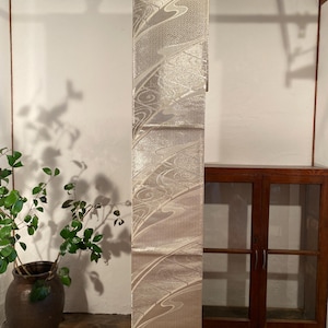 May include: A decorative, cream-colored obi sash with a flowing, wave-like pattern. The sash is hanging vertically from a bamboo rod. The design features silver accents and a subtle texture, creating an elegant aesthetic.