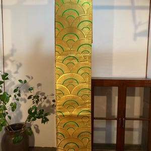 Può includere: Un tessuto dorato con un motivo ripetuto a forma di ventaglio in verde. Il tessuto è appeso verticalmente, mostrando l'intricato design. Il tessuto è probabilmente un oggetto decorativo, forse un runner o un arazzo.