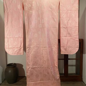May include: A light pink kimono with a floral pattern and red trim. The traditional Japanese garment is hanging, showcasing its long sleeves and intricate design. The fabric has a subtle sheen, and the floral details are evenly spaced.