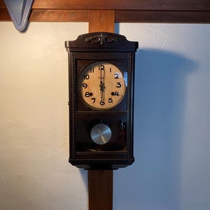 May include: A dark brown antique wall clock with a light-coloured face and black numerals. The clock has a pendulum and is mounted on a wooden beam. The clock is rectangular with a decorative top.