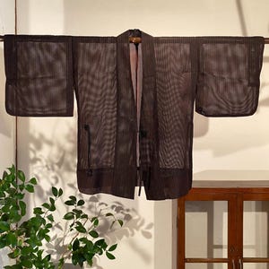 以下が含まれることがあります： 木製の棒に掛けられた、メッシュのような質感のダークブラウンのオープンフロントの着物風ジャケット。 ジャケットは幅広の袖と襟が付いています。 背景には、鉢植えの植物と木製のキャビネットが含まれています。