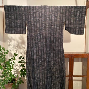 May include: A dark gray kimono with a repeating pattern of vertical, stylized shapes in white and red. The kimono is hanging on a wooden rod, with a plant in a brown pot visible in the background.