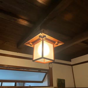 Könnte beinhalten: Eine quadratische Holz-Pendelleuchte mit einer Milchglasscheibe. Das Licht hängt an einer dunklen Holzdecke. Die Leuchte hat einen warmen, einladenden Schein und ist ein dekoratives Element im Raum.