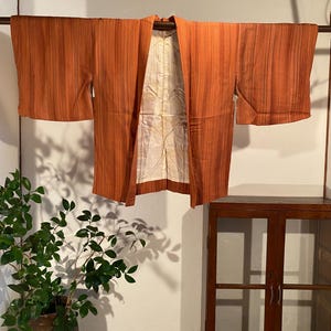 May include: An orange kimono with vertical stripes hangs on a wooden rod. The interior lining is a cream color with a subtle pattern. The garment is displayed against a white wall, with a potted plant and a wooden cabinet in the background.
