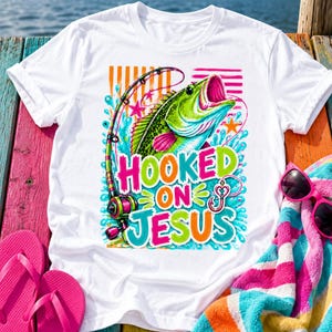 May include: White t-shirt with a colorful graphic of a fish jumping out of the water with a fishing rod. The text "HOOKED ON JESUS" is displayed in bright colors. Pink flip-flops and sunglasses are next to the shirt.