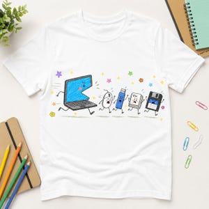 Könnte beinhalten: Ein weißes Kurzarm-T-Shirt mit einer bunten Grafik, die einen blauen Laptop zeigt, der eine Computermaus, einen USB-Stick, eine Esc-Taste und eine Diskette jagt. Die Zeichentrickfiguren haben ausdrucksstarke Gesichter und kleine bunte Sterne.