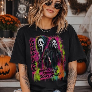Puede incluir: Una camiseta negra con un estampado gráfico colorido del personaje Ghostface sosteniendo un cuchillo y un teléfono. El diseño incluye el texto "GHOSTFACE" y "WHAT'S YOUR FAVORITE SCARY MOVIE?" con vibrantes salpicaduras de pintura neón en verde, rosa y azul.