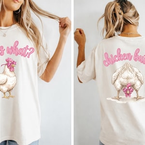 May include: Off-white t-shirt with a whimsical chicken design. The front features the text "guess what?" above a chicken illustration. The back displays "chicken butt" above a chicken illustration. The text and chicken comb are pink.