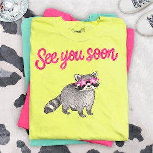 Puede incluir: Una camiseta amarilla neón con la frase "See you soon" en rosa y un mapache con gafas de sol en forma de estrella. La camiseta está superpuesta con camisetas rosas y turquesas.