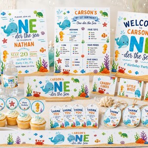 May include: A collection of ocean-themed first birthday party supplies including invitations, milestone signs, welcome signs, water bottle labels, cupcake toppers, food tent cards, and thank you tags featuring whales, crabs, and sea life.