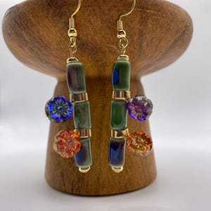 May include: Dangle earrings with gold-coloured hooks. Each earring has a vertical arrangement of green and blue cylindrical beads, separated by gold-coloured spacers. Flower-shaped beads in blue, purple, and orange add colour.
