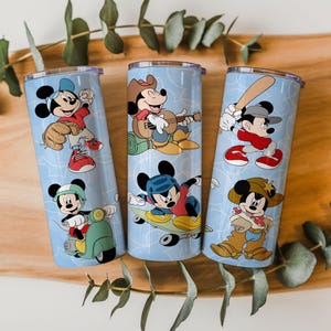 May include: Three cylindrical stainless steel tumblers with a light blue background featuring a repeating pattern of Mickey Mouse in various activities including baseball, playing guitar, riding a scooter, skateboarding, and wearing a cowboy outfit.
