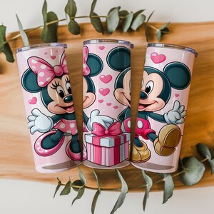 May include: Three cylindrical tumblers with a light pink background featuring cartoon illustrations of Minnie and Mickey Mouse. The design includes small pink hearts, a gift box with a pink bow, and the characters waving, all in a vibrant color palette.