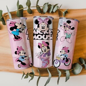 May include: Three cylindrical stainless steel tumblers with clear lids, featuring a light pink background and various illustrations of Minnie Mouse in different outfits and activities, including the text Mickey Mouse printed in black on the center cup.