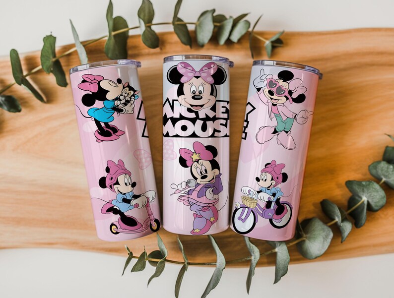 May include: Three cylindrical stainless steel tumblers with clear lids, featuring a light pink background and various illustrations of Minnie Mouse in different outfits and activities, including the text Mickey Mouse printed in black on the center cup.