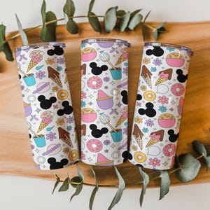 May include: Three cylindrical tumblers with a white background featuring a repeating pattern of black mouse ear silhouettes, colorful ice cream cones, pink donuts, purple bowls, and small flowers. Each tumbler has a clear lid and a smooth finish.