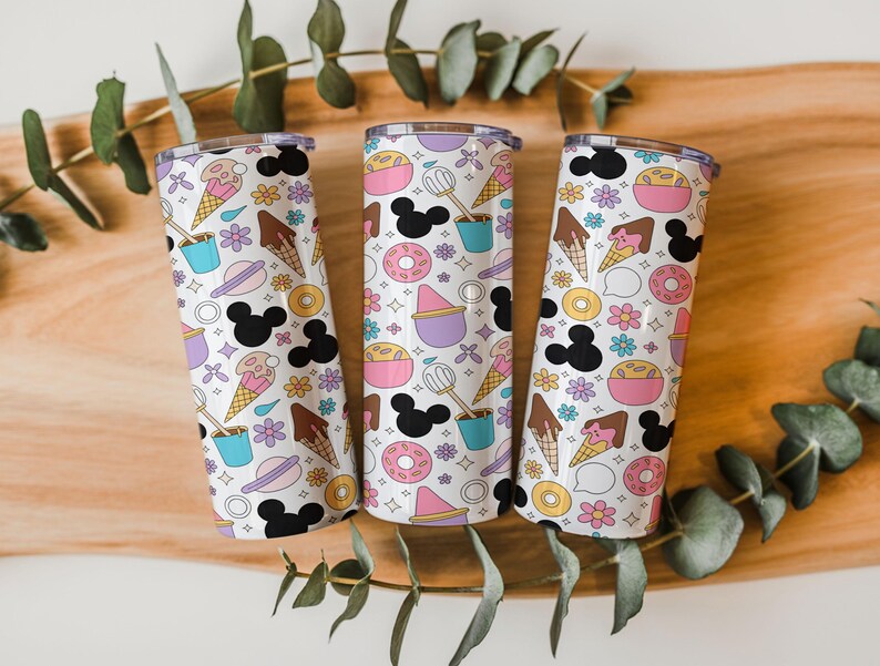 May include: Three cylindrical tumblers with a white background featuring a repeating pattern of black mouse ear silhouettes, colorful ice cream cones, pink donuts, purple bowls, and small flowers. Each tumbler has a clear lid and a smooth finish.