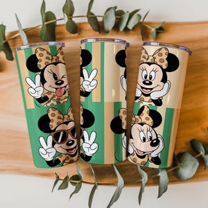 May include: Three cylindrical tumblers featuring a repeating pattern of Minnie Mouse characters. The design includes Minnie with a polka dot bow, making peace signs, wearing sunglasses, and winking, set against a background of vertical green and tan stripes.