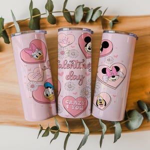 May include: Three light pink cylindrical tumblers with lids, featuring Disney character graphics including Mickey, Minnie, Donald, and Daisy Duck inside pink hearts. The design includes white daisies, small hearts, and text reading Valentine's day, Be Kind, Hug Me, and Crazy 4 You.
