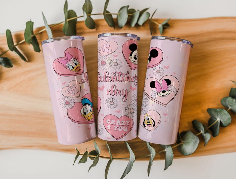May include: Three light pink cylindrical tumblers with lids, featuring Disney character graphics including Mickey, Minnie, Donald, and Daisy Duck inside pink hearts. The design includes white daisies, small hearts, and text reading Valentine's day, Be Kind, Hug Me, and Crazy 4 You.