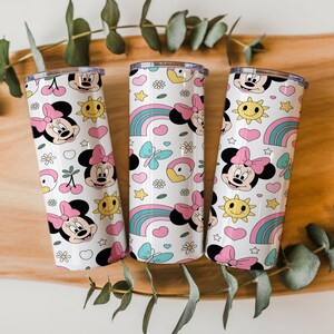 May include: Three cylindrical tumblers with a white background featuring a repeating pattern of Minnie Mouse faces with pink bows, yellow suns, rainbows, blue butterflies, white daisies, pink hearts, and red cherries with green stems.