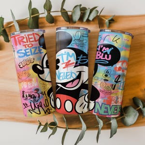 May include: Three cylindrical tumblers featuring colorful graffiti-style artwork with Mickey Mouse imagery. The designs include text such as TRIED TO SEIZE, I'M BLU, and NEVER, layered over vibrant, multi-colored patterns with various cartoon character sketches.