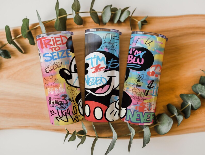 May include: Three cylindrical tumblers featuring colorful graffiti-style artwork with Mickey Mouse imagery. The designs include text such as TRIED TO SEIZE, I'M BLU, and NEVER, layered over vibrant, multi-colored patterns with various cartoon character sketches.