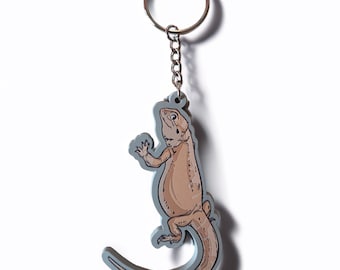 May include: A keychain featuring a cartoon lizard design. The lizard is light brown with a gray outline and a silver key ring. The lizard is in a standing position with its arms raised. The keychain is on a white background.