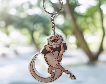 May include: A keychain featuring a cartoon illustration of a bearded dragon lizard. The lizard is tan and brown, with a happy expression and is holding a log. The keychain has a silver chain and ring.