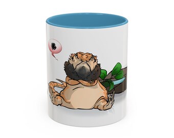 Puede incluir: Una taza de cerámica blanca con interior azul presenta una ilustración de dibujos animados de un dragón barbudo. El dragón es de color canela y marrón con barba negra, y una burbuja de pensamiento está sobre su cabeza. Se ven hojas verdes.