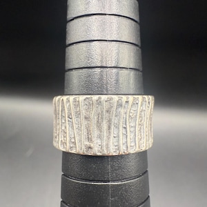 May include: A silver-colored ring with a textured, bark-like design. The ring is wide and sits on a black ring sizer. The ring's surface has vertical lines, creating a unique, organic look. The background is black.