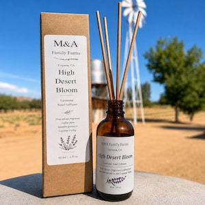 May include: A brown glass bottle of M&A Family Farms High Desert Bloom lavender reed diffuser with four wooden reeds, accompanied by a matching tall rectangular kraft paper box. Both items feature white labels with black text and lavender graphics.