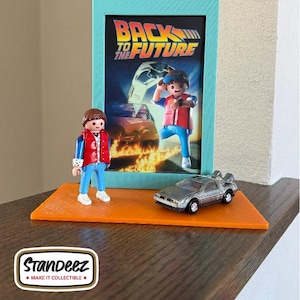 May include: A Back to the Future themed display featuring a framed movie poster, a miniature DeLorean car, and a figure of Marty McFly. The display is set on an orange base with the "Standeez" logo.